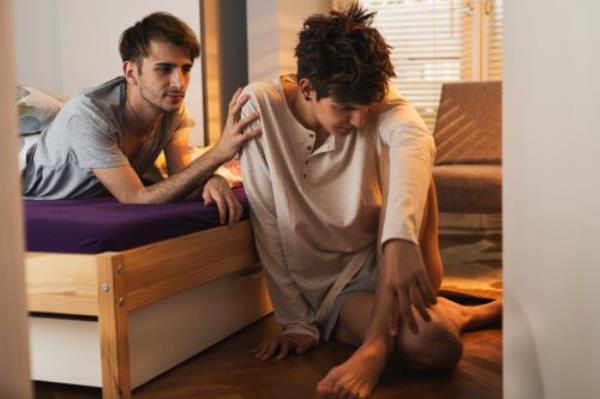 young men on bed putting his arm out to comfort sad man sitting on floor