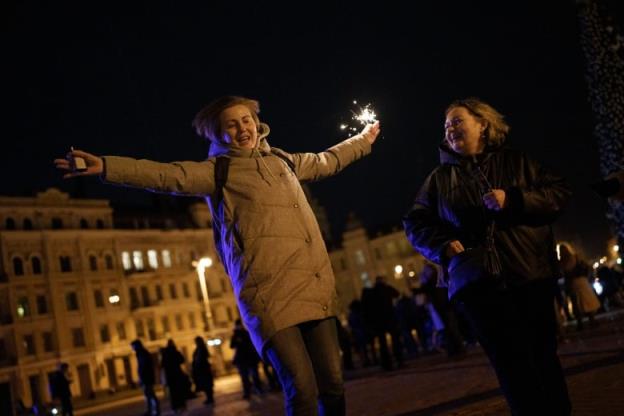 The woman with the sparkler has her arms spread wide. An older woman who looks similar watches, smiling. A small group of people can be seen silhouetted in the distance behind them, with a partially illuminated cityscape beyond.