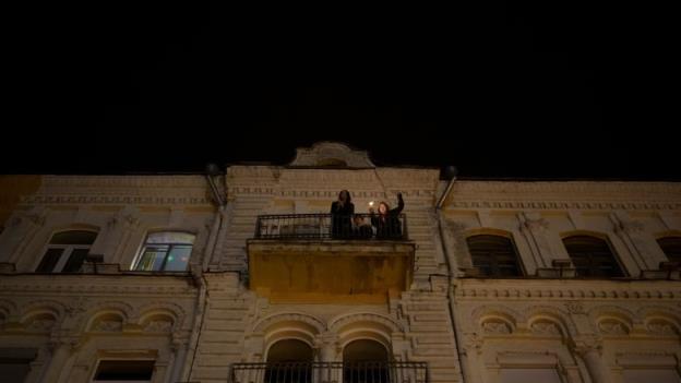 The building is photographed from a point of view on the ground, close to the building. One window has lights on inside, others are dark. Three people are on a railed, stone balcony. One has a light source, possibly a lighter, in their raised hand.